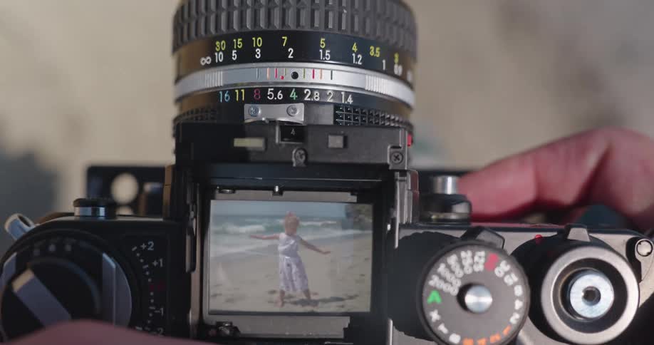A photographer looks through the viewfinder of a vintage SLR camera, focusing on a little girl dancing and waving on a sunny beach