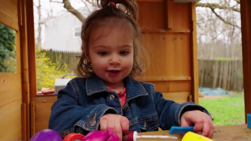 A cute toddler girl joyfully pretends to cut colorful toy vegetables inside her wooden backyard playhouse