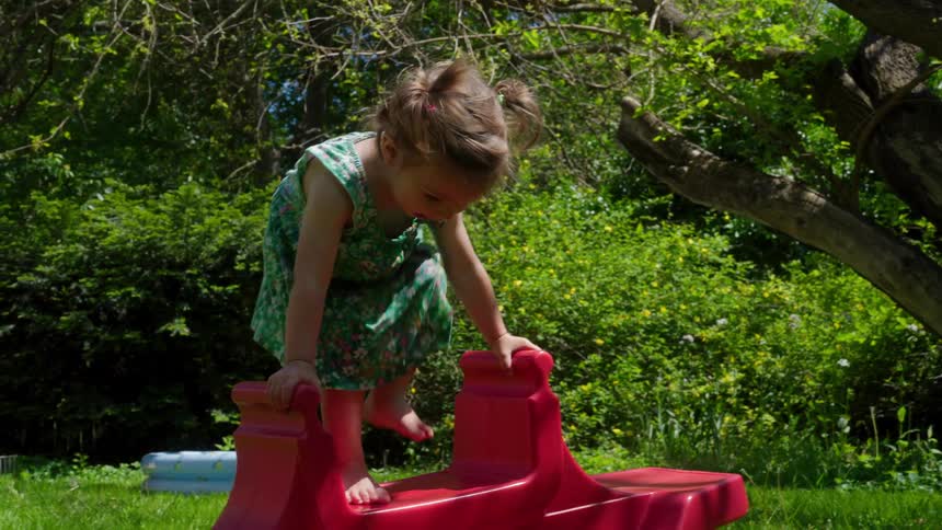 An adorable little girl with pigtails carefully navigates climbing onto a red plastic seesaw in a lush green garden