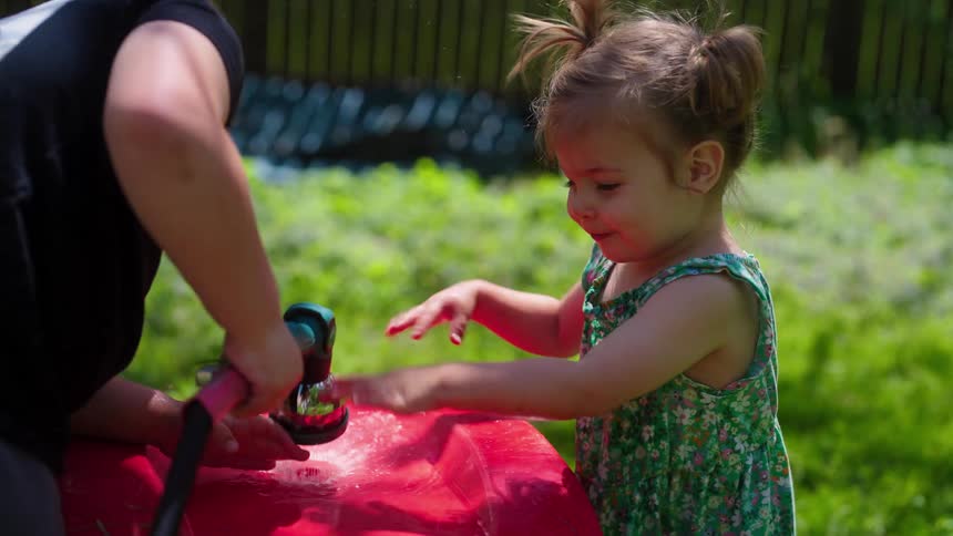 Toddler girl playfully touches water from a garden hose while little boy brother rinses a yard toy in a sunny backyard