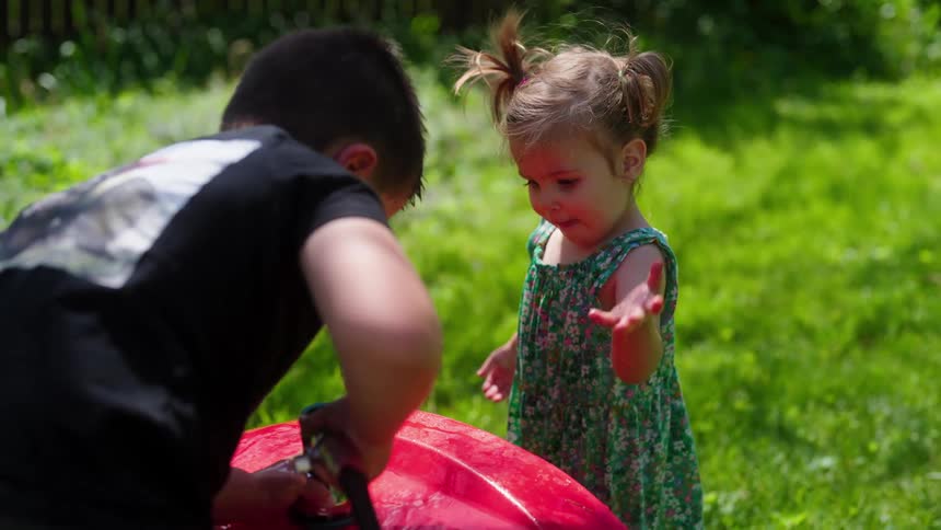 A cute toddler girl with pigtails happily plays with water from a hose held by her brother in a sunny backyard in slow motion