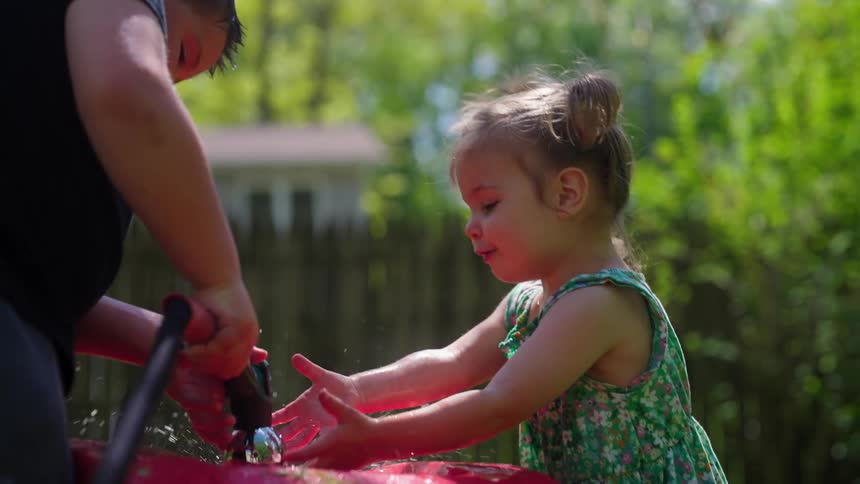A little girl in a floral dress smiles as she plays in the water from a garden hose held by her brother in their backyard