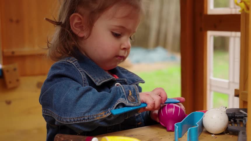 A focused toddler girl in a denim jacket plays with toy food inside her backyard playhouse in slow motion