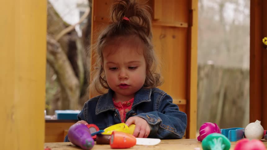 An adorable toddler girl in a denim jacket plays with colorful toy vegetables in a wooden playhouse in the backyard