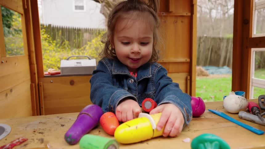 A cute toddler girl happily plays with colorful toy vegetables in her wooden playhouse in the backyard