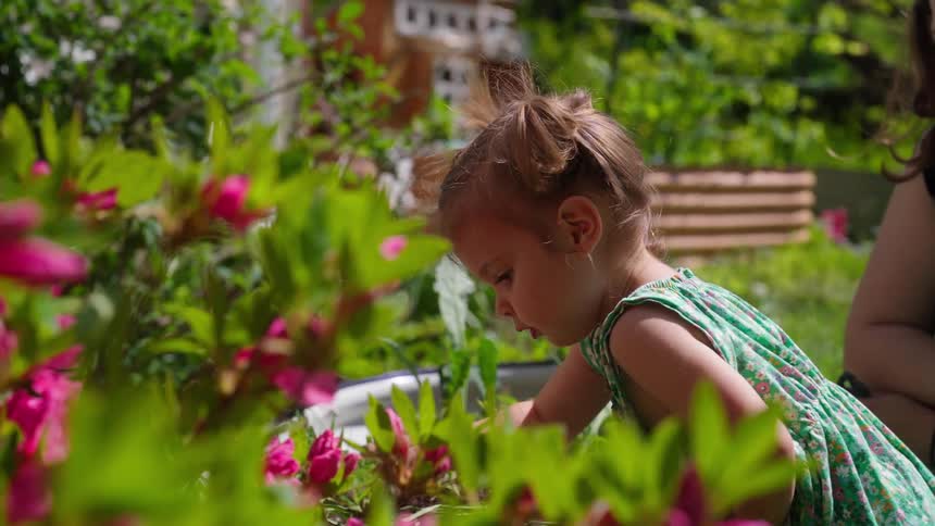 A curious toddler in a green dress plays in a lush, sunny garden while her mother watches lovingly