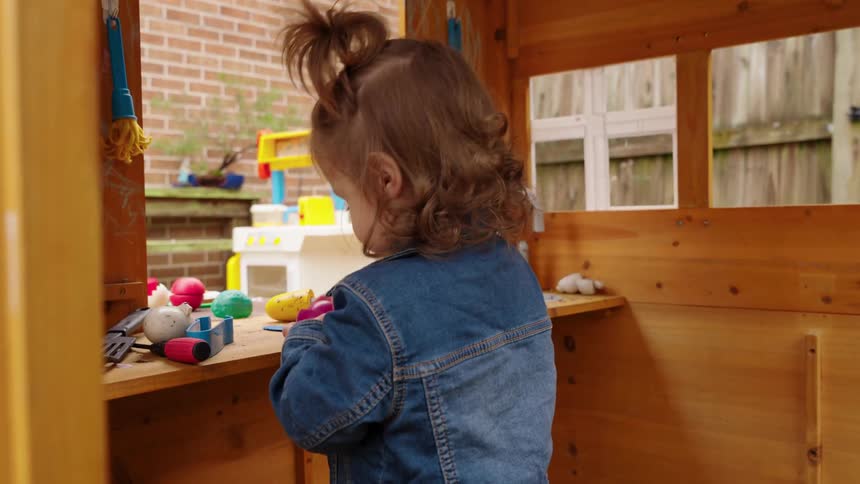 An adorable toddler girl in a denim jacket and yellow boots smiles while playing with her toys in a wooden backyard playhouse