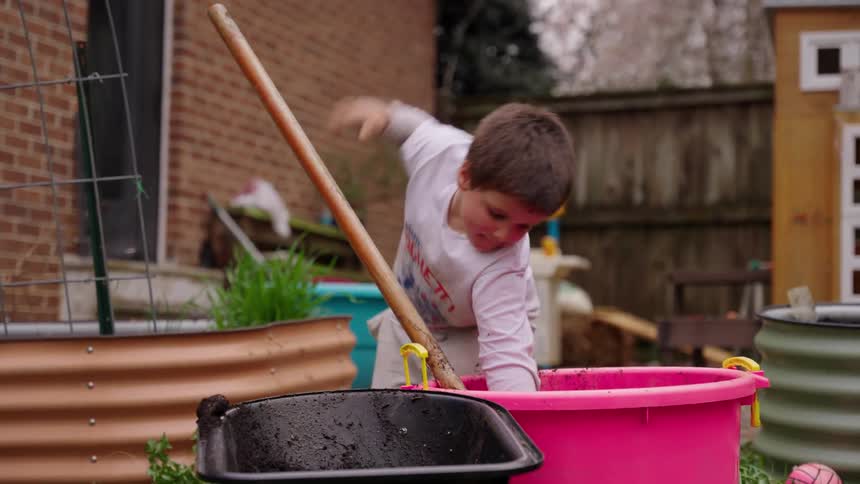 A determined little boy uses a large shovel to move dirt in a backyard garden while his toddler sister watches in slow motion