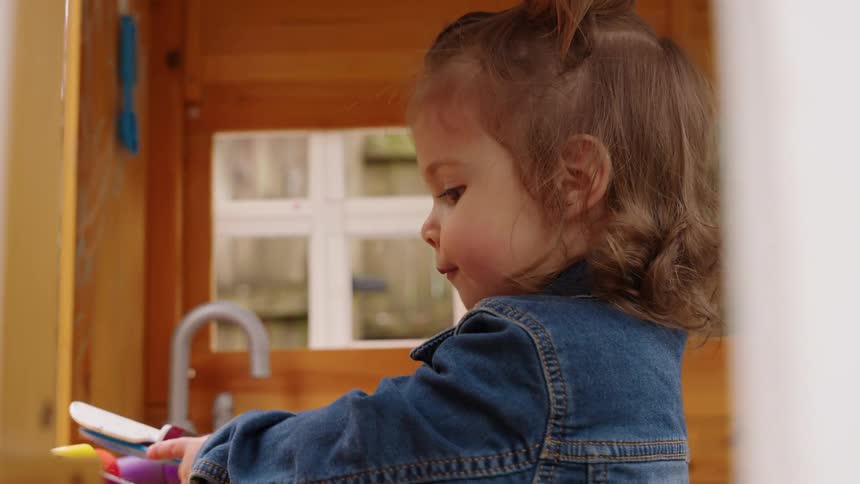 A cute toddler girl in a jean jacket plays happily with her toys in her backyard playhouse in slow motion
