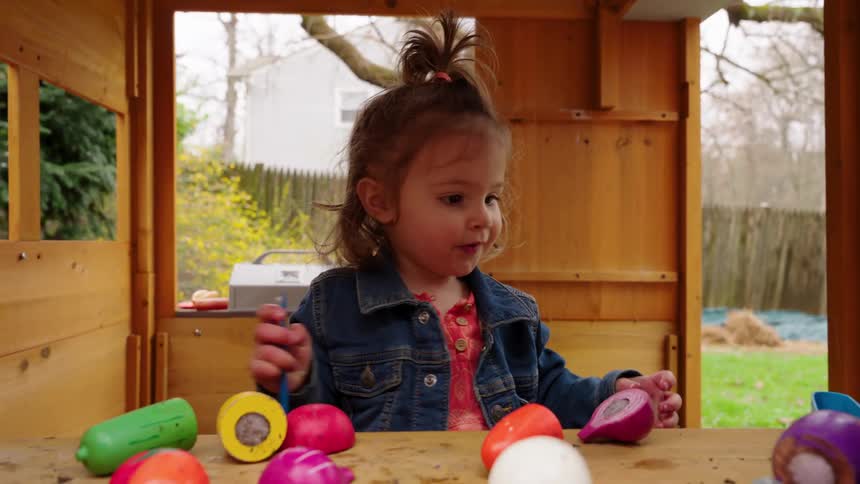 An adorable toddler girl in a denim jacket plays with colorful toy food in a wooden playhouse and then smiles at the camera in slow motion