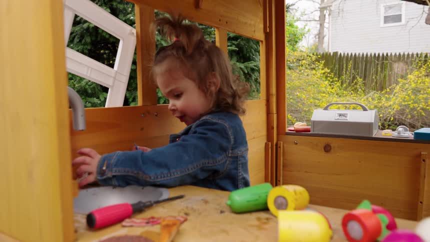 A cute toddler wearing a jean jacket plays with toy vegetables in her wooden playhouse in a slow-motion push-in shot