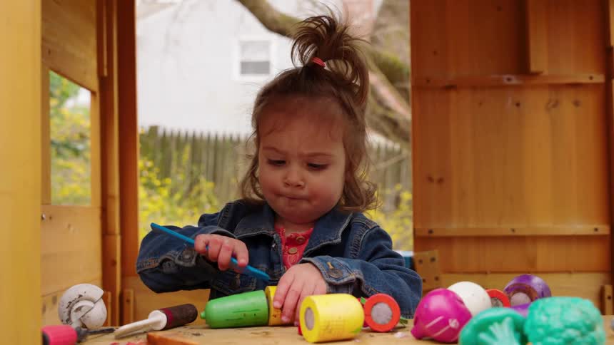 An adorable toddler girl in a denim jacket happily plays with toy food inside a wooden playhouse in the backyard