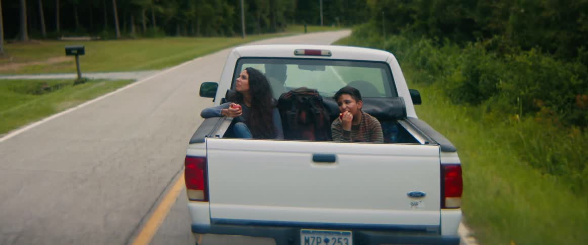 A young woman and a boy eat apples while riding in the back of a pickup truck as it drives down a rural road