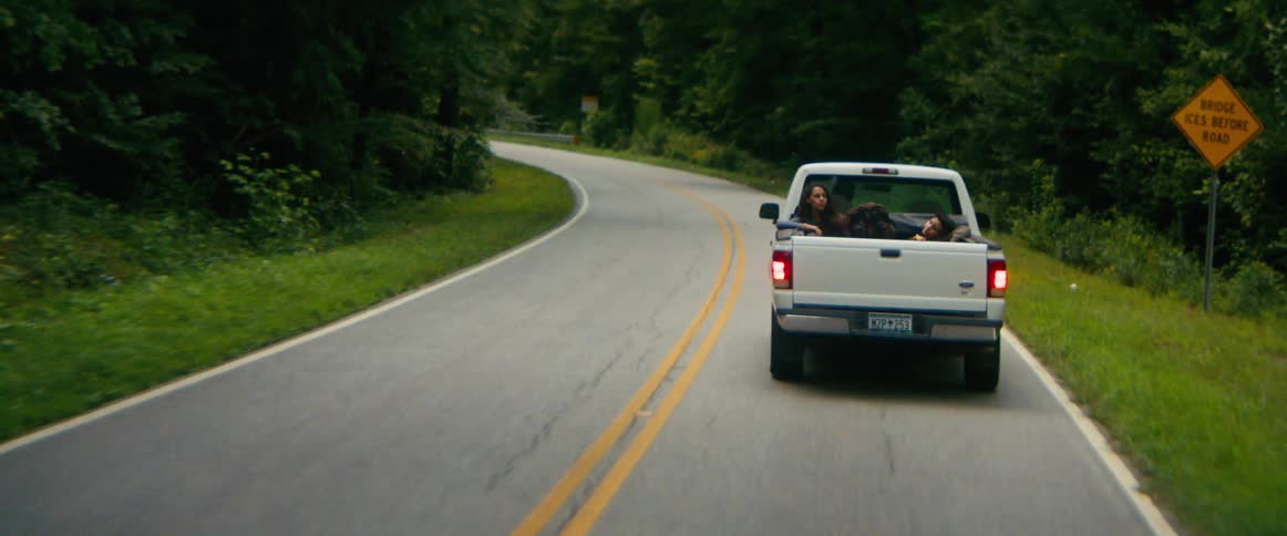 Two young people relax in the bed of a pickup truck as it drives down a winding country road through a lush forest