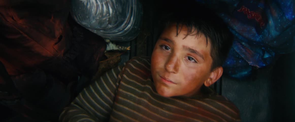 A weary young boy with a dirty face wakes up while traveling in the dark, cramped space of a vehicle