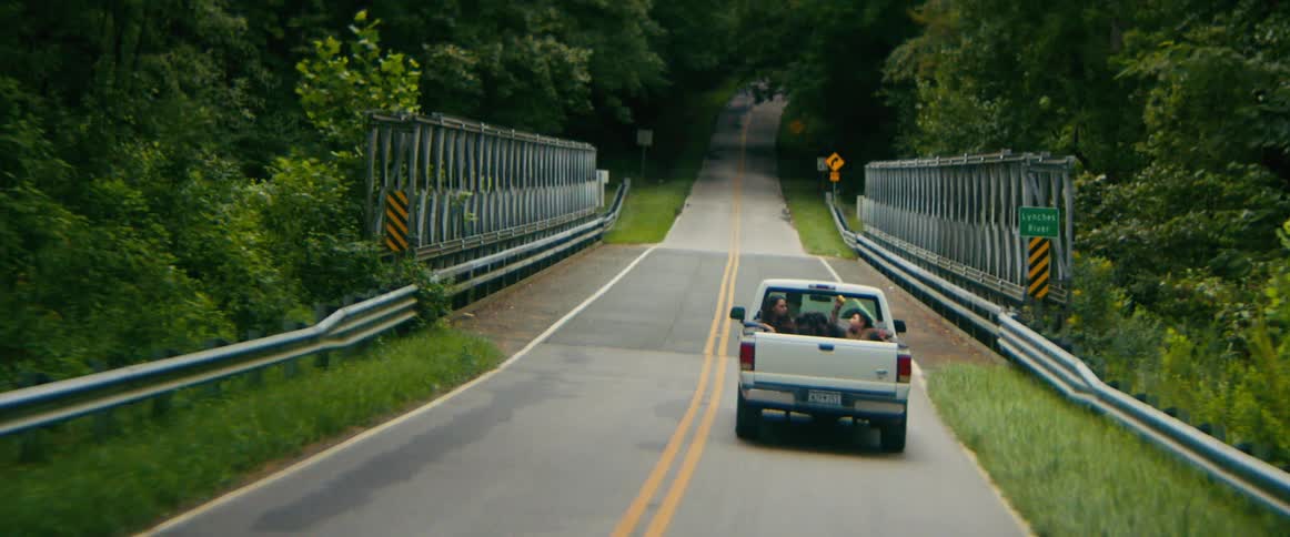 A white pickup truck with friends riding in the back drives away from the camera down a winding, tree-lined country road and across a bridge