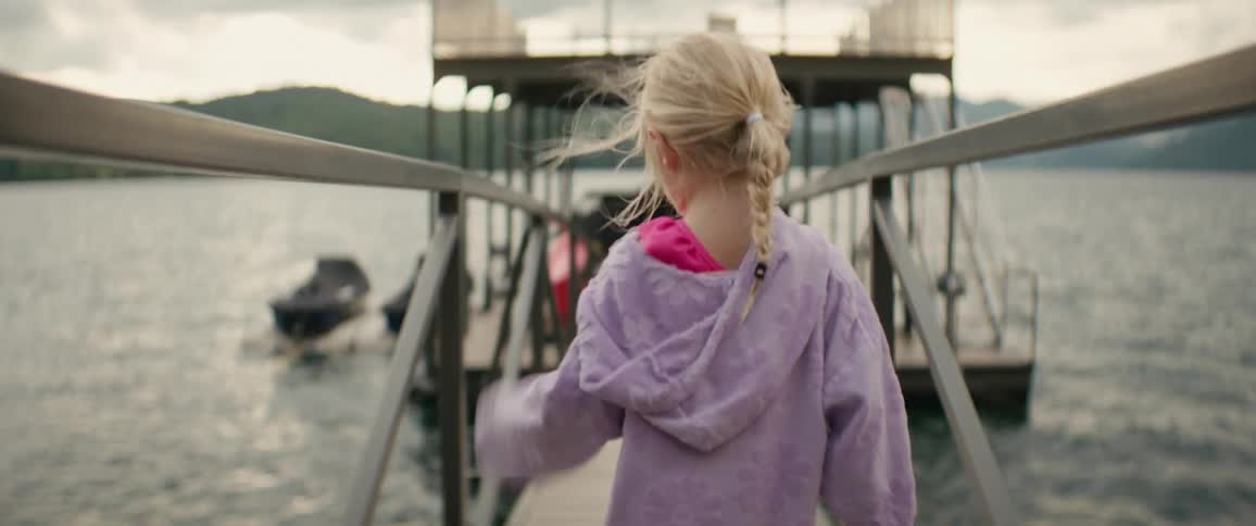 A young girl with braided blonde hair walks down a floating dock ramp towards the boats on a lake