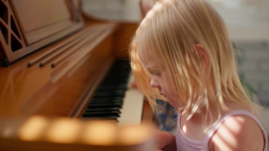 Two young blonde sisters concentrate while playing a wooden piano together in a sunlit room