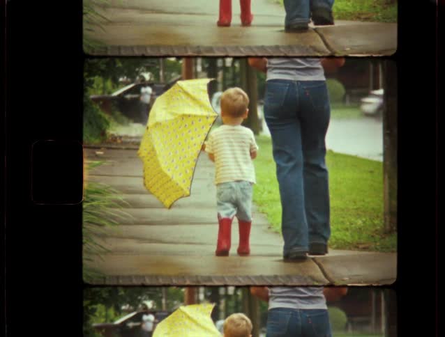 A young boy in red rain boots holding a yellow umbrella walks with his mother down a wet sidewalk in this vintage-style clip