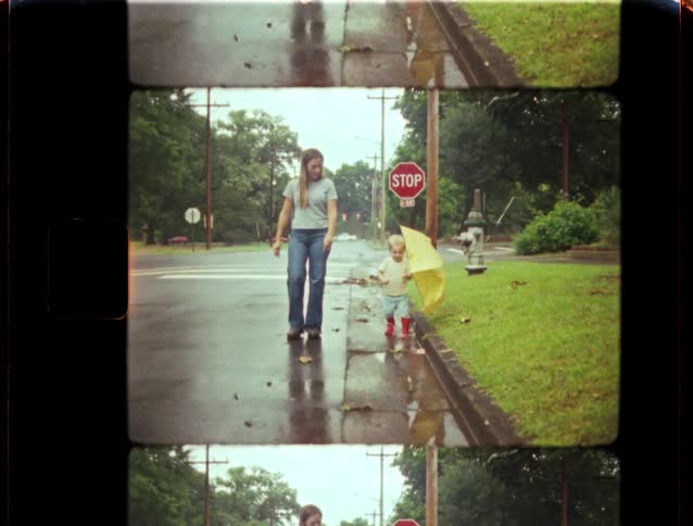 A woman and a small child holding a yellow umbrella walk down a wet sidewalk on a rainy day in this nostalgic 8mm film scan