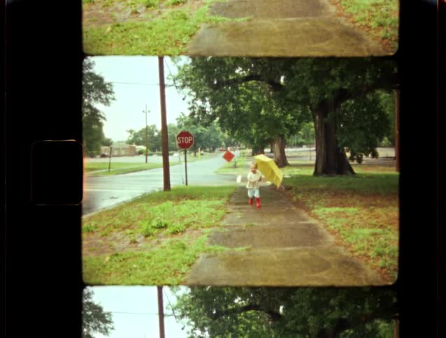 A cute toddler with a big yellow umbrella and bright red rain boots playfully runs down a wet sidewalk on a rainy day
