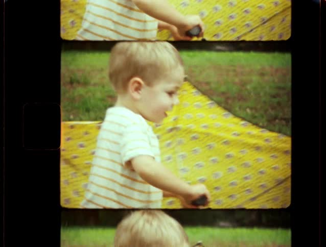 A nostalgic home movie shows a toddler playing with a yellow patterned umbrella in the backyard