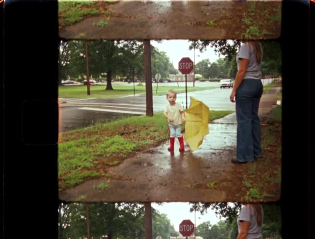 A nostalgic home movie shows a toddler in bright red rain boots holding a yellow umbrella while standing in the rain with their mother