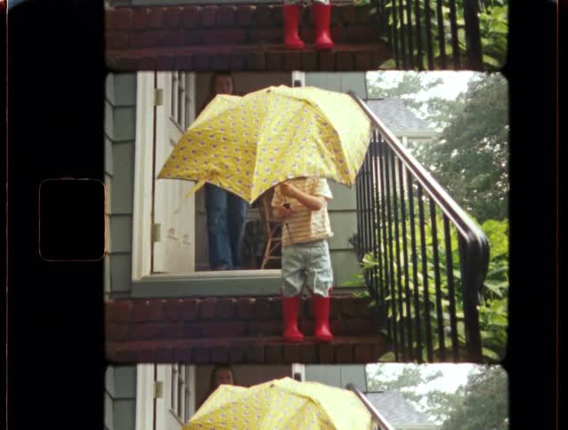 Vintage film footage shows a mother watching her toddler in red rain boots play with a yellow umbrella on their front steps