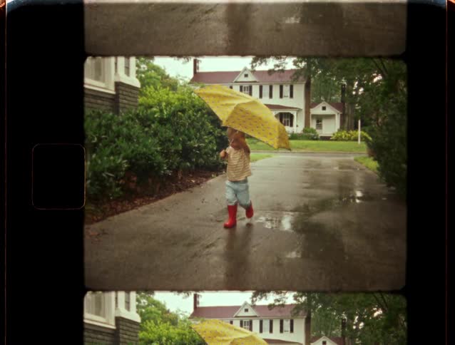 Vintage home movie captures a cute toddler in red rain boots running towards the camera with a big yellow umbrella on a rainy day