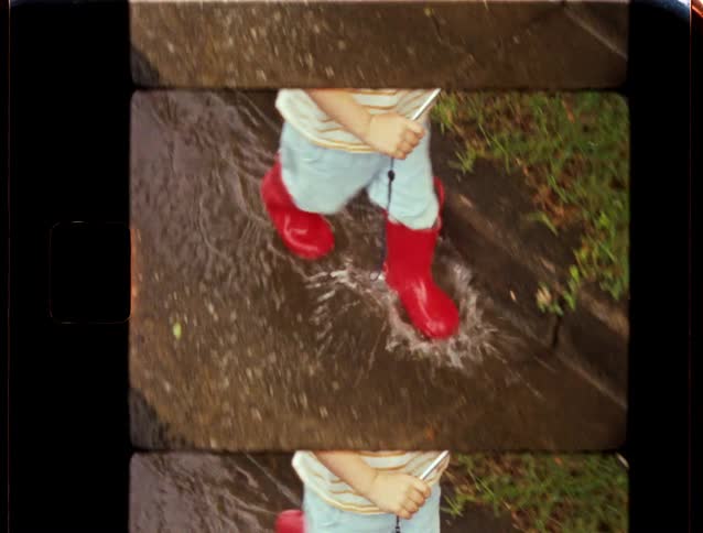 Vintage 8mm home movie of a toddler's bright red rain boots splashing in a puddle on the sidewalk