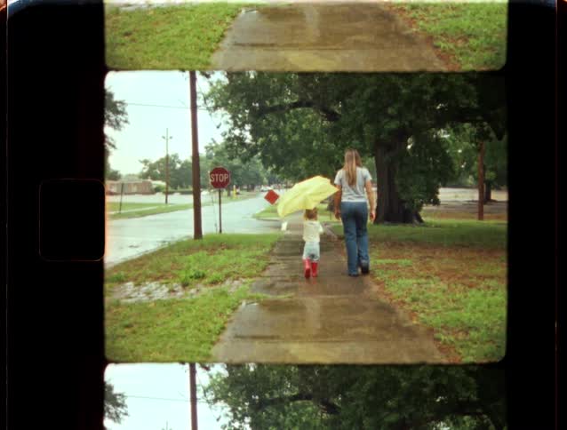 A mother and her young child walk down a sidewalk on a rainy day with a yellow umbrella in this vintage 8mm home movie