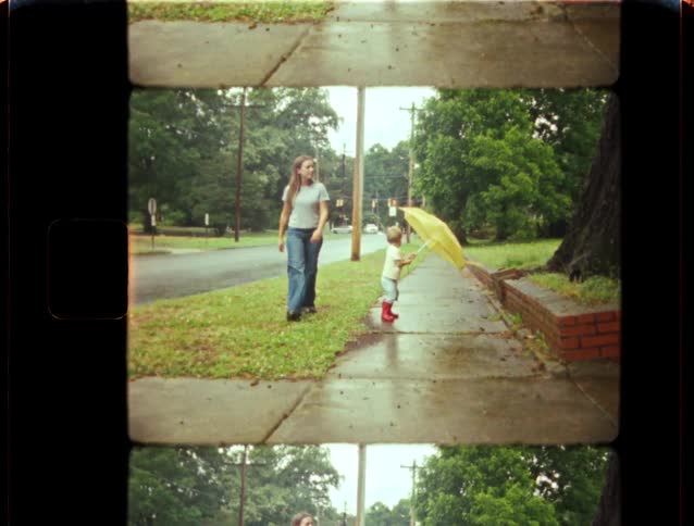 A mother and her young child in red rain boots walk with a yellow umbrella on a rainy day in this vintage 8mm film clip