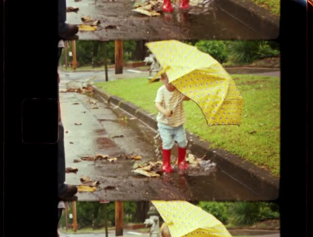 A little boy in bright red rain boots splashes in a puddle on the street while holding a big yellow umbrella in this charming archival home movie footage