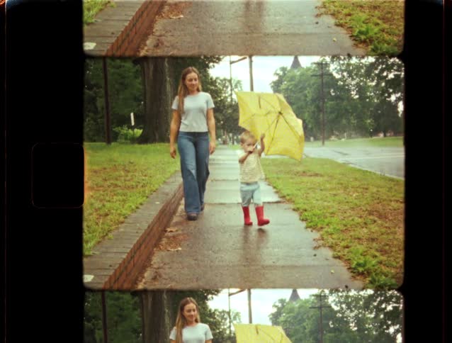 A young boy in red rain boots walks with his mother under a yellow umbrella on a rainy day in this charming vintage footage