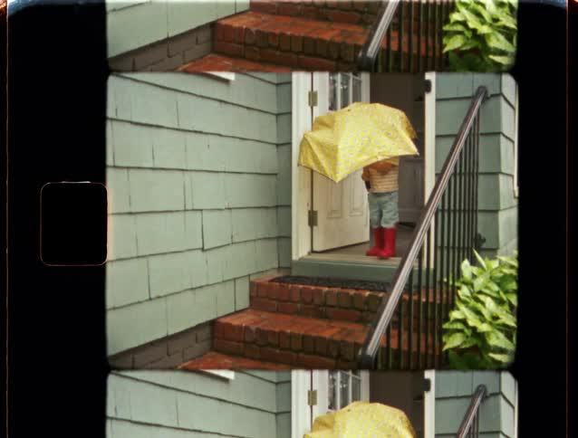 Vintage 8mm film captures a toddler in red rain boots playfully opening a large yellow umbrella in a doorway