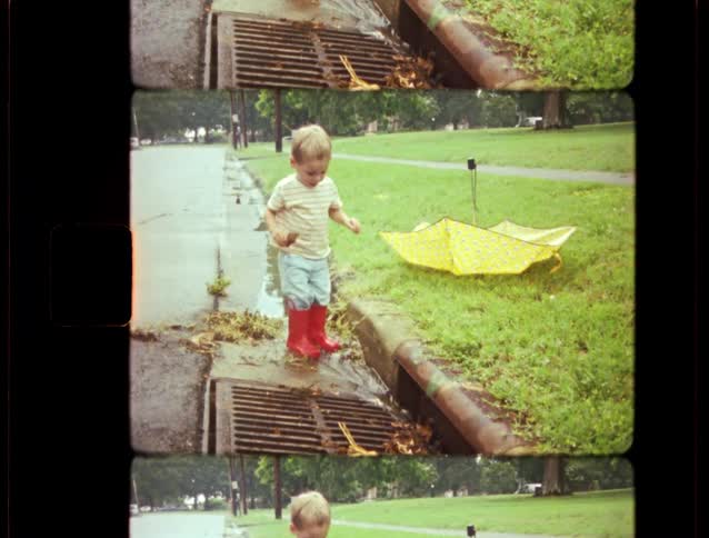 A toddler in bright red rain boots stands by a storm drain watching the water flow after a rainstorm in this vintage home movie clip