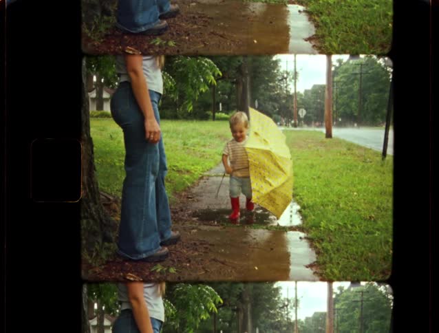 Vintage footage shows a happy toddler with an umbrella and red boots splashing in puddles with his mother on a rainy day