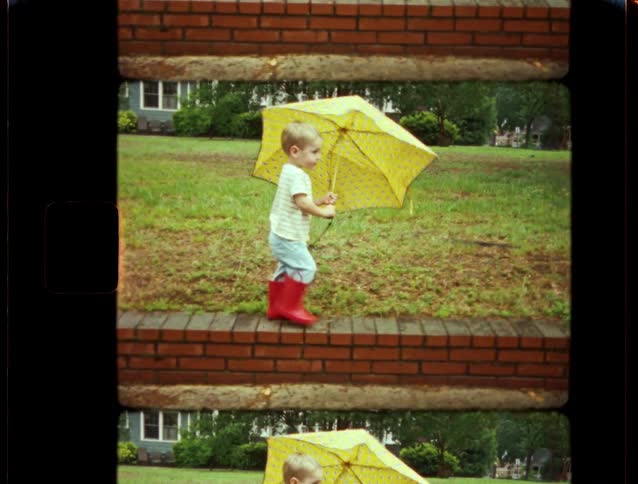 Charming vintage film captures a little boy in bright red rain boots walking on a brick wall with a big yellow umbrella