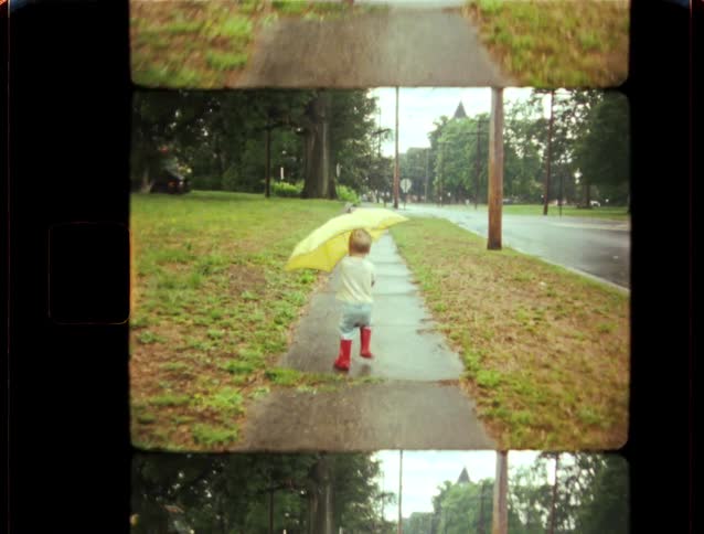 Vintage home movie footage shows a little boy in red rain boots walking with a big yellow umbrella on a sidewalk after a rainstorm