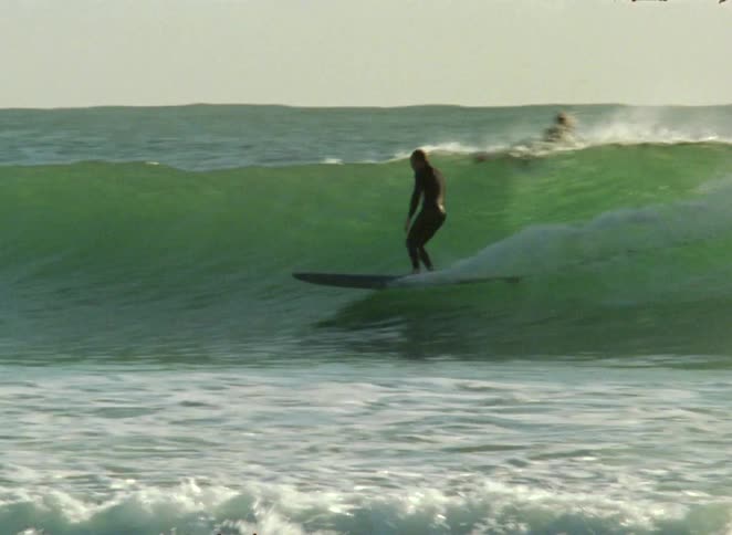 A surfer in a full wetsuit glides smoothly across a green ocean wave on a longboard