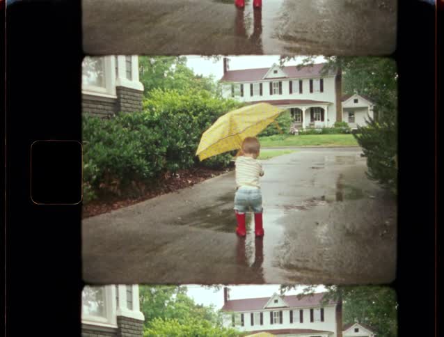 A nostalgic home movie captures a young boy in bright red rain boots holding a yellow umbrella in the rain