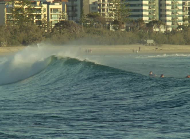 A surfer catches a breaking wave sending up a large spray while other surfers wait near a city beach