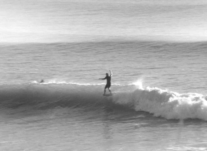Archival black and white footage shows a surfer riding a wave and then wiping out in the ocean