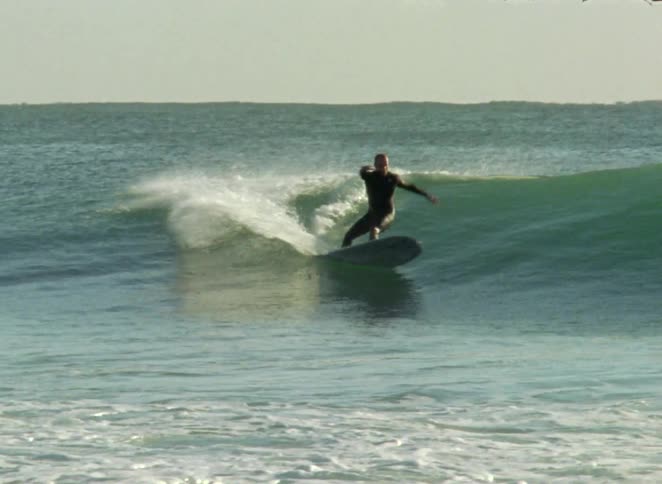 A surfer in a wetsuit gracefully carves across a beautiful green ocean wave on a longboard in slow motion