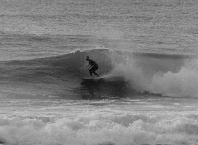 A lone surfer in a wetsuit successfully rides out of the barrel of a huge ocean wave in black and white