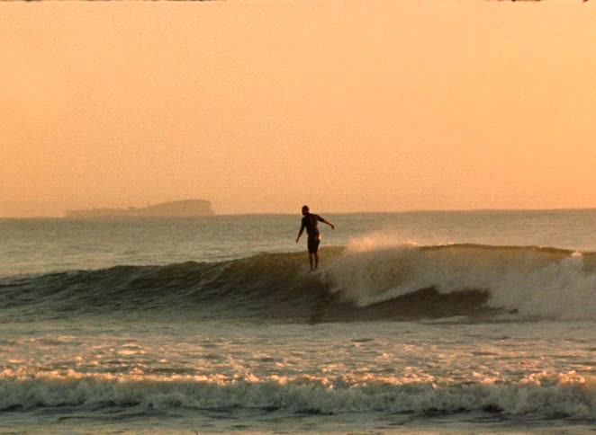 A silhouetted surfer glides across a wave in slow motion against a golden sky at sunset