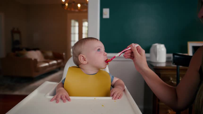 A loving mother spoon-feeds her adorable baby boy who sits happily in his high chair at home