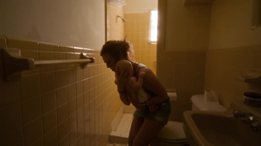 A distressed mother gently dries her baby on the floor of a dimly lit, rundown bathroom
