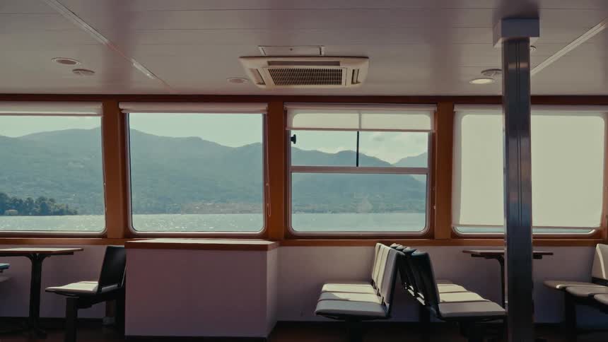 A serene view of sunlit water and green mountains from the empty passenger cabin of a ferry boat