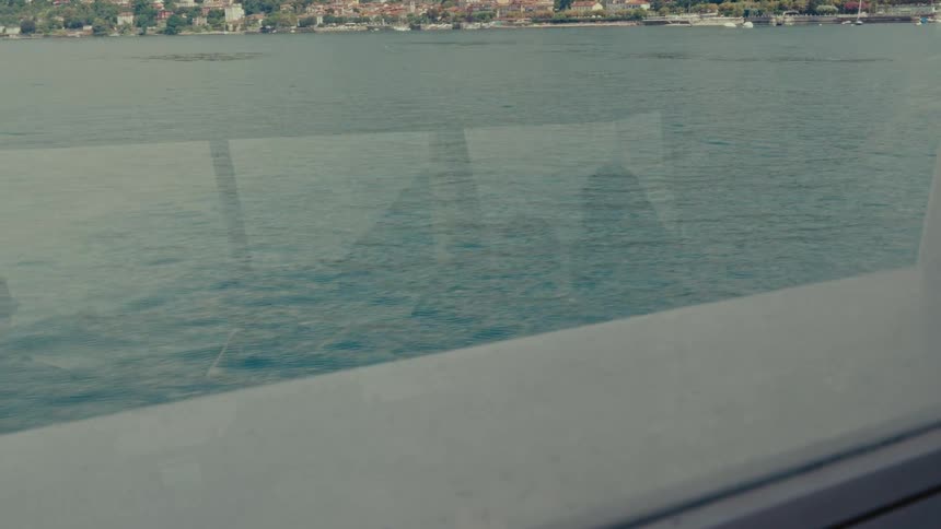 A scenic view of a coastal town across the water from the window of a moving boat, with passenger reflections in the glass